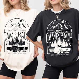 May include: Two t-shirts with a black and white graphic design of mountains, trees, a tent, and a campfire. The text on the shirts reads "JUNE 2024 Ava's Bachelorette CAMP BACH BRECKENRIDGE, CO". One shirt is ivory and the other is black.