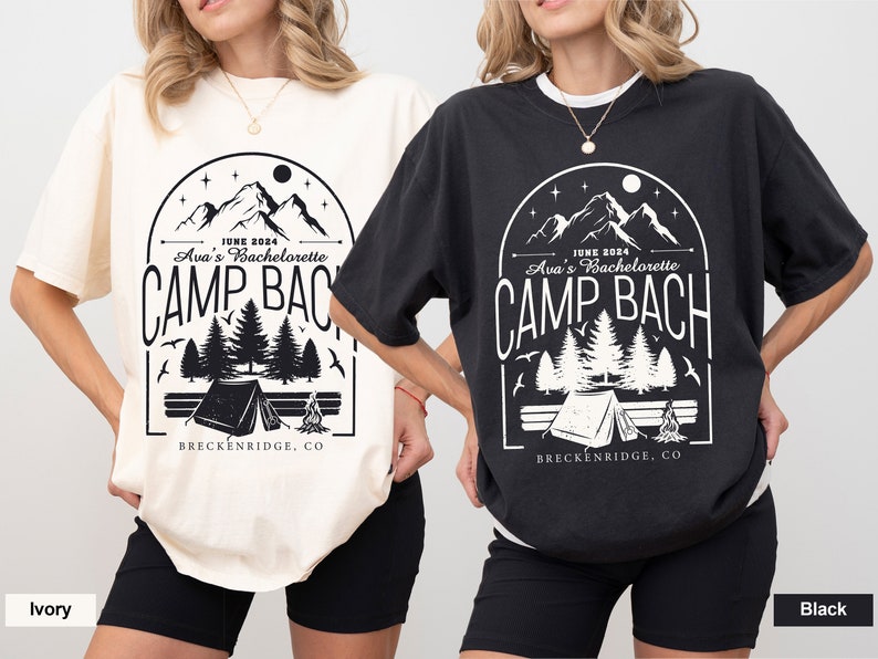 May include: Two t-shirts with a black and white graphic design of mountains, trees, a tent, and a campfire. The text on the shirts reads "JUNE 2024 Ava's Bachelorette CAMP BACH BRECKENRIDGE, CO". One shirt is ivory and the other is black.