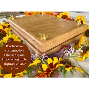 May include: A rectangular wooden flower press featuring two thick boards held together by four metal bolts with brass wing nuts. A circular brown graphic overlay contains the text: Reach out for customization! Choose a quote, image, or logo to be engraved on your press.