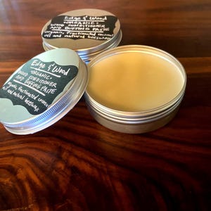 May include: Two silver tins of Edge & Wood organic wood conditioner and buffing paste. The labels are black with white text. The tins are on a dark brown wooden surface.
