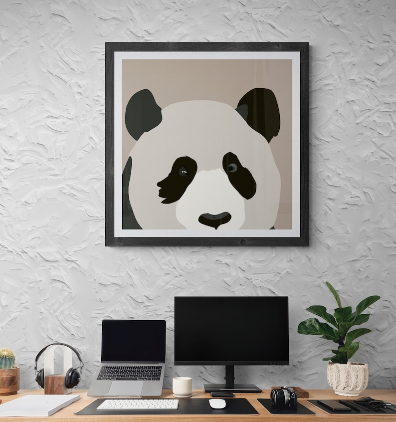 May include: A framed illustration of a panda bear's face with black and white fur. The panda is looking directly at the viewer. The frame is black and the background is a light brown color. The image is hanging on a white wall above a desk with a computer, laptop, and other office supplies.