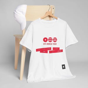 Könnte beinhalten: Weißes T-Shirt mit roter Grafik mit der Aufschrift "BTS WORLD TOUR". Das Shirt hat einen Rundhalsausschnitt und kurze Ärmel. Eine weiße Baseballkappe liegt auf der Rückseite eines Holzstuhls.