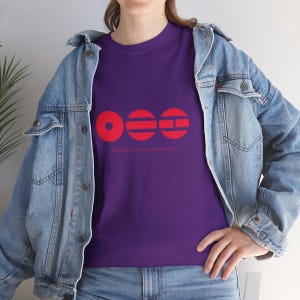Könnte beinhalten: Ein lila T-Shirt mit einem roten Grafikdesign und dem Text "YOU KNOW THAT BTS IS BACK!". Die Grafik besteht aus drei roten Formen: einem Kreis, zwei horizontalen Linien und einem Rechteck. Das Shirt ist aus weichem Material.