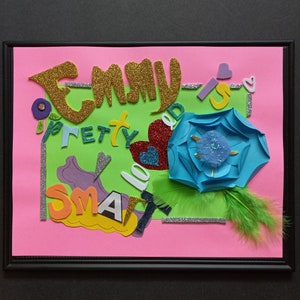 Può includere: Un'opera d'arte incorniciata con sfondo rosa e verde. L'opera presenta il nome "Emmy" in lettere glitterate dorate, insieme alle parole "pretty", "smart" e "loved". Sono inclusi anche un fiore di carta blu e piume verdi.