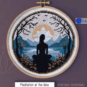 Pode incluir: Padrão de ponto cruz de uma pessoa meditando à beira de um lago com montanhas ao fundo. O padrão está em um aro de bordado de madeira e inclui 8 cores DMC e 110 x 112 pontos.
