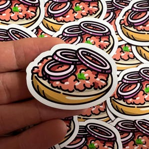 Könnte beinhalten: Ein Set bunter Sticker mit einer Cartoon-Illustration eines deutschen Mettbrötchens mit roten Zwiebelringen darauf. Die Sticker sind auf weißem Papier gedruckt.