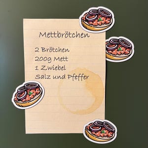 Könnte beinhalten: Ein handgeschriebenes Rezept für Mettbrötchen, ein deutsches Gericht, auf liniertes Papier, mit Cartoon-Aufklebern des Gerichts. Das Rezept enthält Zutaten wie 2 Brötchen, 200g Mett, 1 Zwiebel, Salz und Pfeffer. Ein Kaffeefleck ist sichtbar.