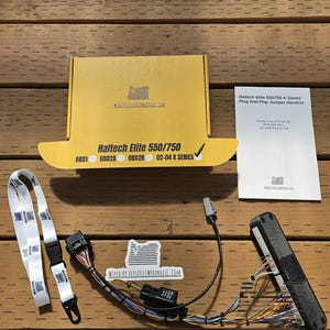 May include: An assortment of automotive wiring components, including a box labeled "Haltech Elite 550/750," a white lanyard, and a wiring harness. The components are arranged on a wooden surface.