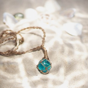 May include: A turquoise glass bead encased in a macrame knot pendant hangs from a brown cord necklace. The necklace is lying on a white sandy surface.