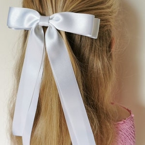 Könnte beinhalten: Eine weiße Haarschleife aus Satin mit langen Bändern ist in blondem Haar befestigt. Die Schleife ist klassisch gestaltet und verleiht einen Hauch von Eleganz. Dieses Haaraccessoire ist eine einfache, aber stilvolle Ergänzung für jede Frisur.