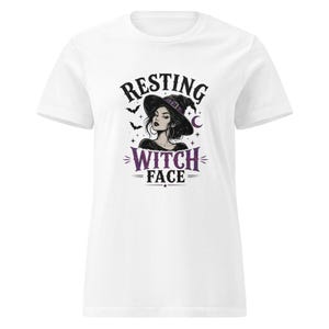 May include: A white short-sleeved t-shirt featuring a graphic print of a person wearing a purple-accented witch hat. The text "RESTING WITCH FACE" is printed in black and purple lettering, accompanied by small bat, star, and crescent moon icons.