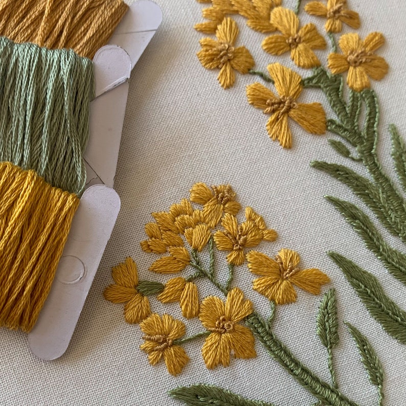 May include: Detailed embroidery of yellow flowers and green leaves on a cream-coloured fabric. Spools of yellow and green embroidery thread are positioned on the left. The floral design highlights the craft's intricate stitching.