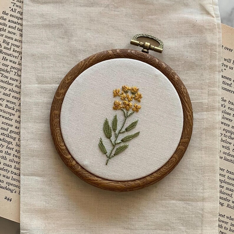 May include: Hand-stitched floral embroidery in a wooden hoop. The design showcases a yellow flower and green leaves on a white fabric. The hoop is approximately 15 cm in diameter, a delicate piece of handmade art.