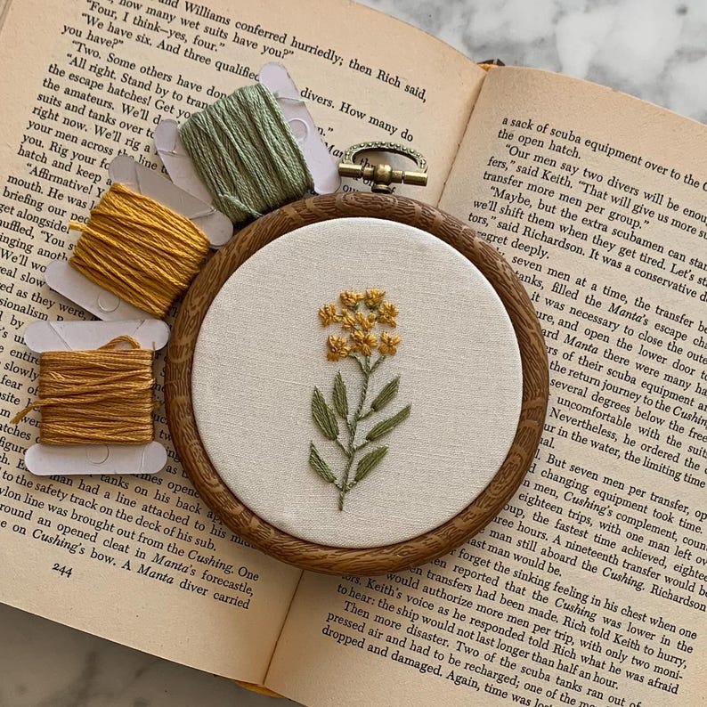 May include: An embroidered floral design with yellow flowers and green leaves, set in a wooden hoop. The image includes spools of yellow and green thread. The background features an open book. The hoop measures approximately 10 cm in diameter.