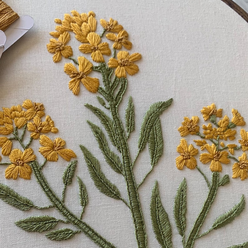 May include: Close-up of an embroidery hoop with a floral design. The embroidery features yellow flowers with detailed stitching and green stems and leaves. The background is a light-coloured fabric, highlighting the vibrant colours of the embroidery.