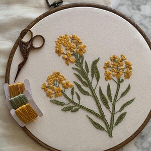 May include: An embroidered floral design on a cream-coloured fabric stretched within a wooden hoop. The design features yellow flowers and green leaves. A pair of small scissors and spools of yellow and green thread are also visible.