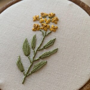 May include: Hand-stitched embroidery art featuring a yellow flower with green leaves and stem. The floral design is centered on a cream-coloured fabric, set within a wooden embroidery hoop. The embroidery showcases detailed thread work.