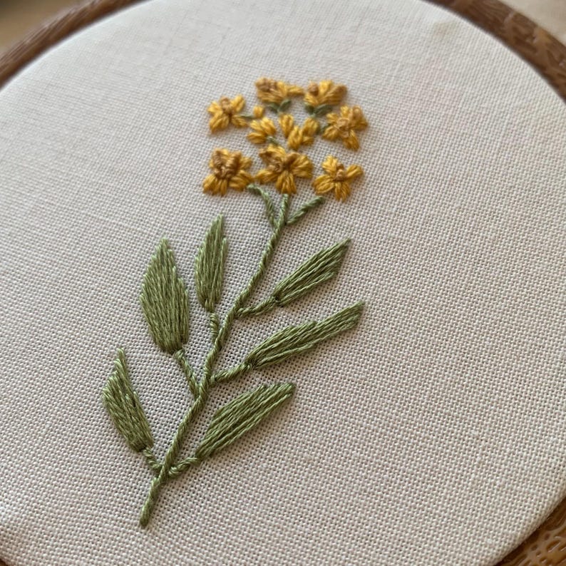 May include: Hand-stitched embroidery art featuring a yellow flower with green leaves and stem. The floral design is centered on a cream-coloured fabric, set within a wooden embroidery hoop. The embroidery showcases detailed thread work.