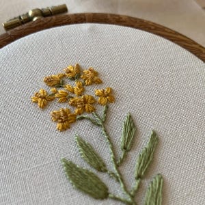 May include: Close-up of an embroidery hoop with a floral design. The artwork features yellow flowers with green leaves and stems, stitched on cream fabric. The wooden hoop is visible. The embroidery is a delicate craft.
