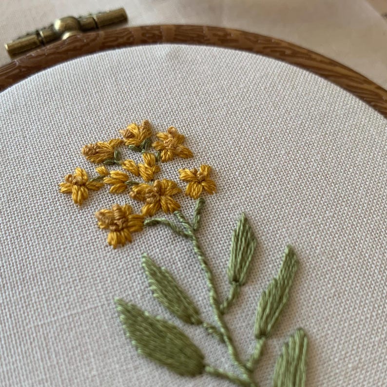 May include: Close-up of an embroidery hoop with a floral design. The artwork features yellow flowers with green leaves and stems, stitched on cream fabric. The wooden hoop is visible. The embroidery is a delicate craft.