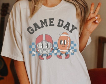 Könnte beinhalten: Ein weißes T-Shirt mit dem Text "GAME DAY" in schwarzen Buchstaben. Das Shirt zeigt einen Cartoon-Footballhelm und einen Football, die sich die Hände halten.