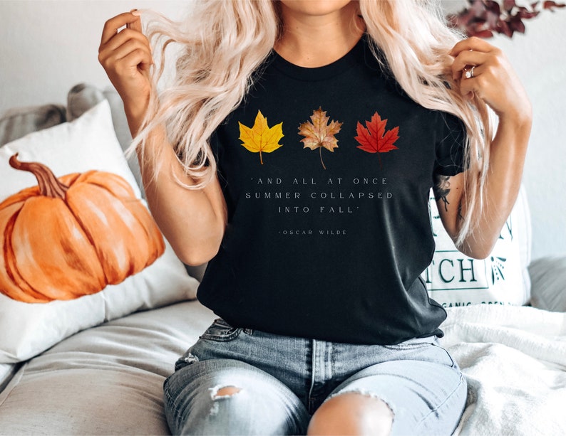 Puede incluir: Camiseta negra con una cita de Oscar Wilde: "Y de repente, el verano se derrumb&oacute; en oto&ntilde;o." La camiseta presenta tres hojas de oto&ntilde;o de colores.