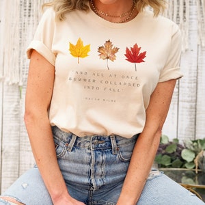 Puede incluir: Una camiseta beige con una cita de Oscar Wilde: "Y de repente, el verano se derrumb&oacute; en oto&ntilde;o." La camiseta presenta tres hojas de oto&ntilde;o en tonos de amarillo, naranja y rojo.