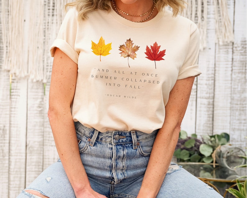 Puede incluir: Una camiseta beige con una cita de Oscar Wilde: "Y de repente, el verano se derrumb&oacute; en oto&ntilde;o." La camiseta presenta tres hojas de oto&ntilde;o en tonos de amarillo, naranja y rojo.