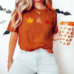 Puede incluir: Una camiseta de color &oacute;xido con una cita de Oscar Wilde sobre el verano que se convierte en oto&ntilde;o. La cita dice "Y de repente el verano se derrumb&oacute; en oto&ntilde;o." La camiseta presenta tres hojas de oto&ntilde;o estilizadas en amarillo, naranja y rojo.