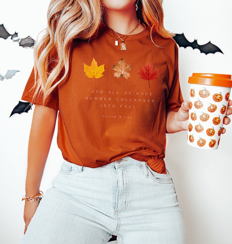 Puede incluir: Una camiseta de color &oacute;xido con una cita de Oscar Wilde sobre el verano que se convierte en oto&ntilde;o. La cita dice "Y de repente el verano se derrumb&oacute; en oto&ntilde;o." La camiseta presenta tres hojas de oto&ntilde;o estilizadas en amarillo, naranja y rojo.