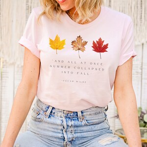 Puede incluir: Una camiseta rosa con una cita de Oscar Wilde: "Y de repente el verano se derrumb&oacute; en oto&ntilde;o." La cita est&aacute; rodeada de tres hojas de oto&ntilde;o en tonos de amarillo, naranja y rojo.