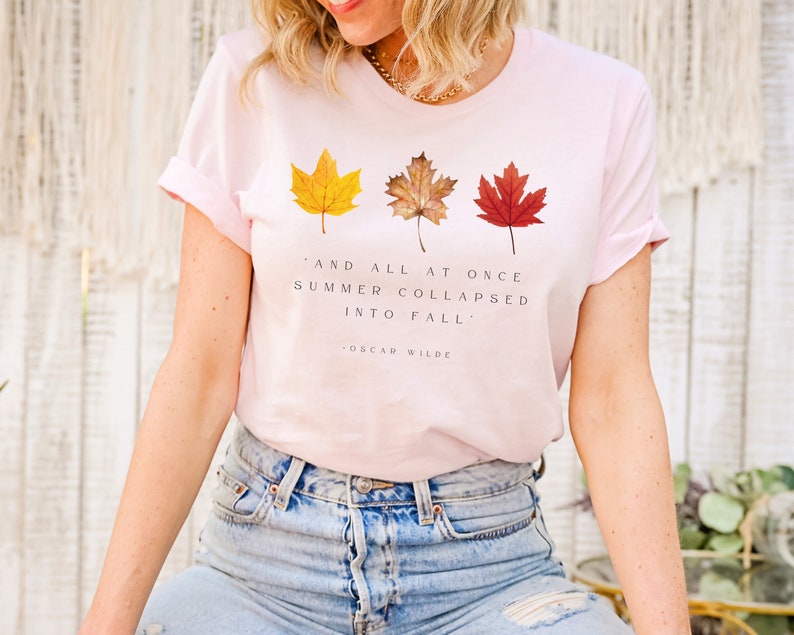 Puede incluir: Una camiseta rosa con una cita de Oscar Wilde: "Y de repente el verano se derrumb&oacute; en oto&ntilde;o." La cita est&aacute; rodeada de tres hojas de oto&ntilde;o en tonos de amarillo, naranja y rojo.