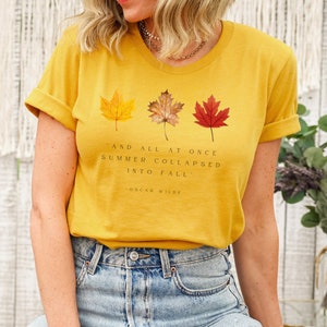 Puede incluir: Una camiseta amarilla mostaza con una cita de Oscar Wilde: "Y de repente, el verano se derrumb&oacute; en oto&ntilde;o." La camiseta presenta tres hojas de oto&ntilde;o en tonos de rojo, naranja y amarillo.