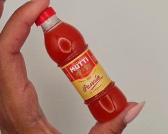 May include: A small, clear plastic bottle filled with red tomato passata. The bottle features a red screw-top cap and a yellow label with red text that reads Mutti, 100% Italiano, and Passata di Pomodoro, featuring a small lion logo graphic.