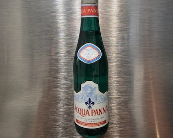 May include: A green glass bottle of Acqua Panna natural spring water with a silver cap. The label features a blue fleur-de-lis emblem, red text reading Acqua Panna, and a small oval crest with blue and red borders. The bottle is filled with clear liquid.