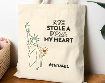 May include: A white canvas tote bag featuring a green outline of the Statue of Liberty holding a slice of pizza. The text "NYC STOLE A PIZZA MY HEART" is printed in black. The name "MICHAEL" is printed in black at the bottom of the bag.