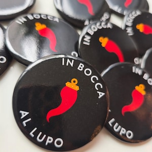 Könnte beinhalten: Schwarze runde Knöpfe mit dem weißen Text "IN BOCCA AL LUPO". Jeder Knopf zeigt eine rote Chilischote mit einer goldenen Krone. Die Knöpfe sind auf einer weißen Oberfläche angeordnet.