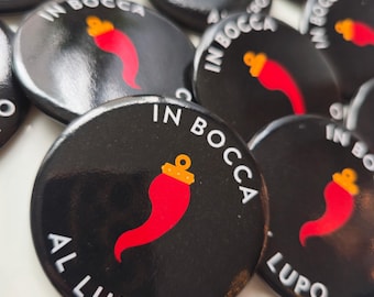 May include: Black circular badges with the Italian phrase "IN BOCCA AL LUPO" in white lettering. Each badge displays a red chili pepper with a gold crown. The badges are arranged on a white surface.