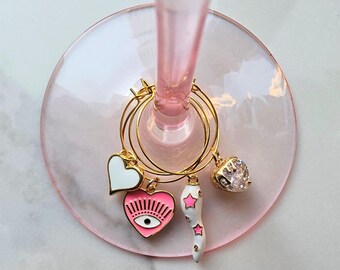 May include: Gold wine glass charms with decorative pendants. Charms include a white heart, a pink heart with an eye design, a white horn with pink stars, and a gold heart with a clear gem. The charms are hanging on a pink wine glass.