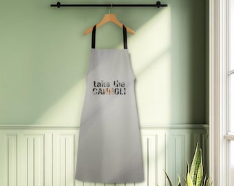 May include: A light grey apron with black straps hangs from a wooden hanger. The apron features the text "take the CANNOLI" in a mix of black, grey, and tan letters. The apron is hanging on a black rod against a light green wall.