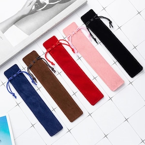 May include: A selection of velvet pen pouches in various colours: blue, brown, red, pink, and black. Each pouch features a drawstring closure. The pouches are arranged diagonally on a white surface.