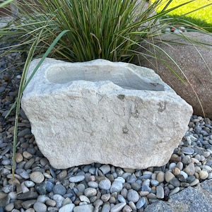 Könnte beinhalten: Ein rechteckiger, hellgrauer Steinpflanzer mit einem ausgehöhlten Becken. Der Pflanzer steht auf einem Bett aus kleinen, glatten Steinen, mit hohem grün-gelbem Gras im Hintergrund. Ein großer, abgerundeter Felsen ist ebenfalls zu sehen.