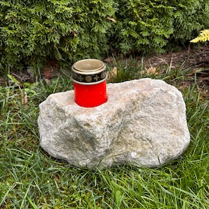 Könnte beinhalten: Ein roter Kerzenhalter mit goldenem Deckel und dekorativen Symbolen sitzt in einem grauen Stein auf einem Bett aus grünem Gras.