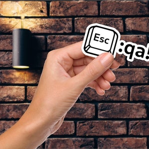 以下が含まれることがあります： 手の中に、黒い枠で囲まれたエスケープキーと「Esc」という文字が書かれた白いステッカーが置かれている。そのステッカーの隣には、「:q a!」という文字が書かれた白いステッカーがある