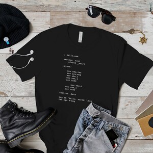 Könnte beinhalten: Schwarzes T-Shirt mit weißem Text, der Assembler-Code zeigt. Der Code enthält die Zeilen: 'section .text', 'global _start', '_start:', 'mov edx, len', 'mov ecx, msg', 'mov eax, 1', 'int 0x80', 'section .data', 'msg db "Hello, world!", 0xa', 'len equ $-msg'.