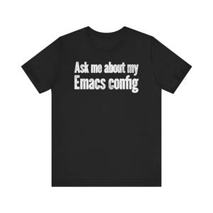 Puede incluir: Una camiseta negra de manga corta con el texto blanco "Ask me about my Emacs config" impreso en el pecho. Las letras tienen un aspecto ligeramente desgastado y texturizado sobre la tela negra sólida de la prenda.