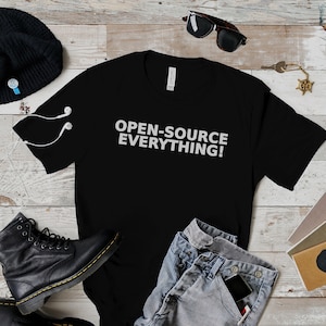 Peut inclure: T-shirt noir avec le texte "OPEN-SOURCE EVERYTHING!" en blanc. Le t-shirt est présenté avec des bottes noires, un jean bleu, un bonnet noir, des lunettes de soleil et des carnets sur une surface en bois.