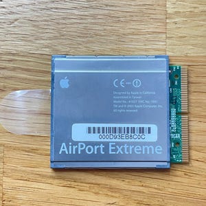 Puede incluir: Una tarjeta AirPort Extreme plateada con el logotipo de Apple, modelo A1027 y el texto "AirPort Extreme". La tarjeta tiene un borde de placa de circuito verde y está sobre una superficie de madera.
