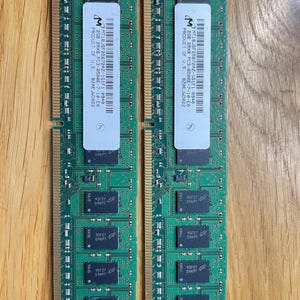 May include: Two green RAM memory modules with gold-colored connectors. Each module has a white label with black text, including "PRODUCT OF U.S." and other technical specifications. The modules are designed for computer use.