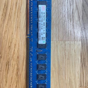 May include: A blue RAM stick with the text "hynix 1GB 1Rx8 PC3-10600E-9-10-D0" and other technical details. The RAM stick is rectangular and has several black chips. It is placed on a wooden surface.
